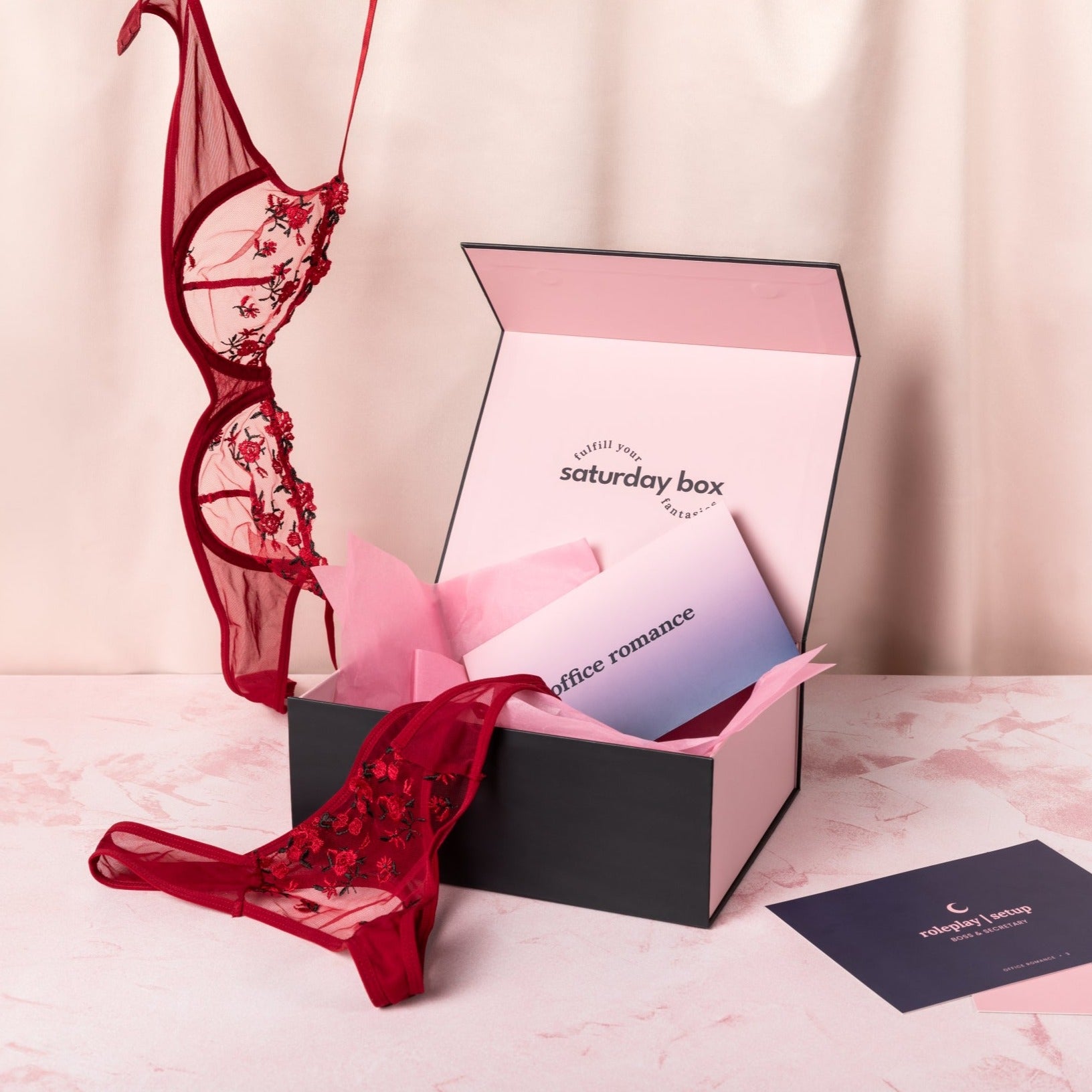 Opened Office Romance Classic Box with pink tissue paper and branded envelope inside with floral lingerie bra dangling from the top of frame and panties draped over the edge of the box. Gameplay cards are in the foreground next to the box.
