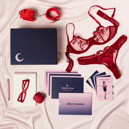 The components of the Office Romance Classic box are all laid out on a silky fabric including the magnetic box, the bright red fuzzy cuffs, the memo pad and pink pen, the bright red secretary glasses, the rose shaped suction vibrator, the red floral lingerie, and an assortment of gameplay and character cards included with the set. 