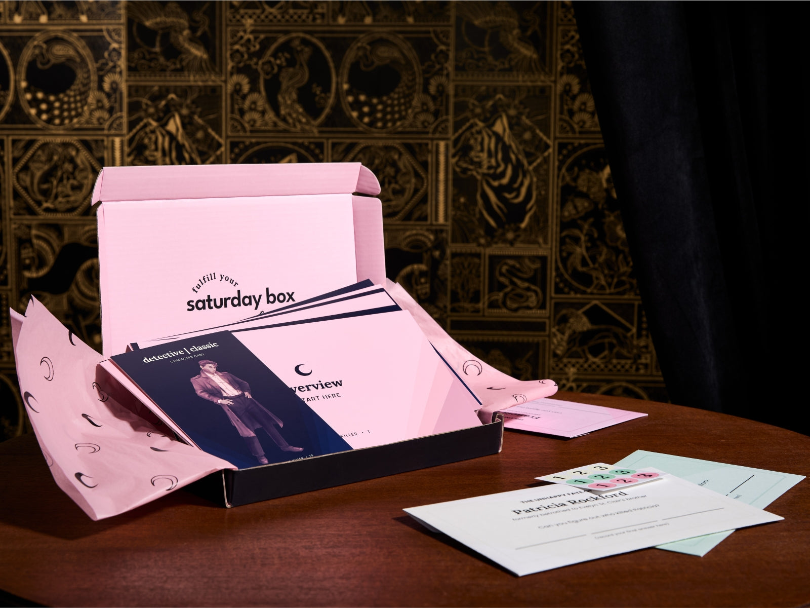 An open pink 'Saturday Box' package sits on a wooden table, showing printed inserts and a few cards in front. The background features ornate dark gold wallpaper and a black curtain.