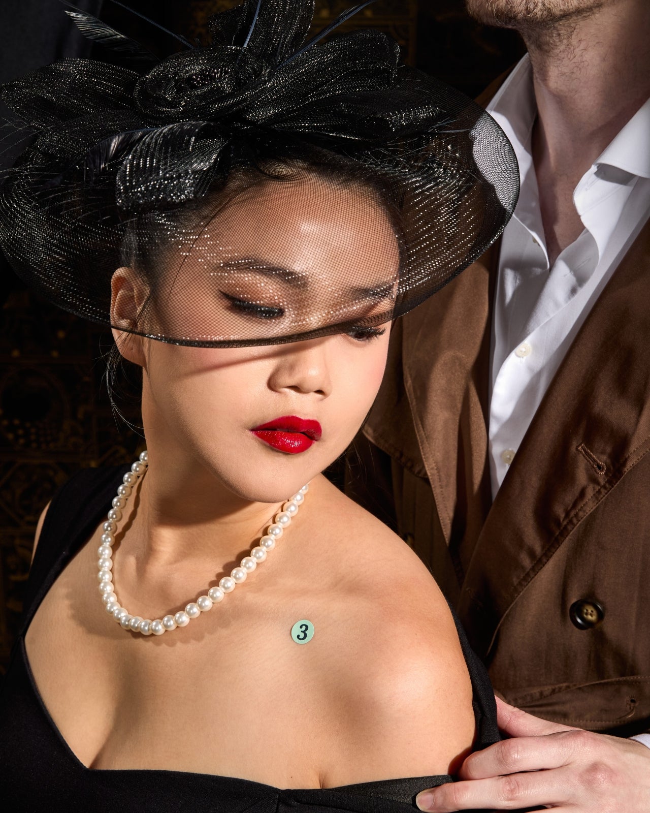 Close-up of the dame wearing a black fascinator, a string of pearls, and the clue sticker on her shoulder.