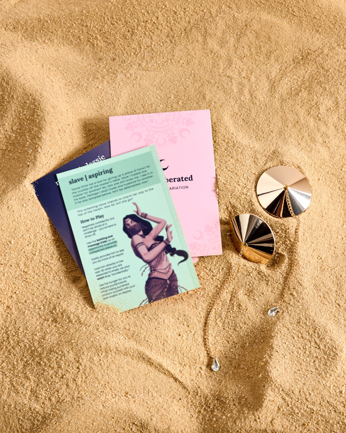 Three cards and two gold jewelry pieces on sand