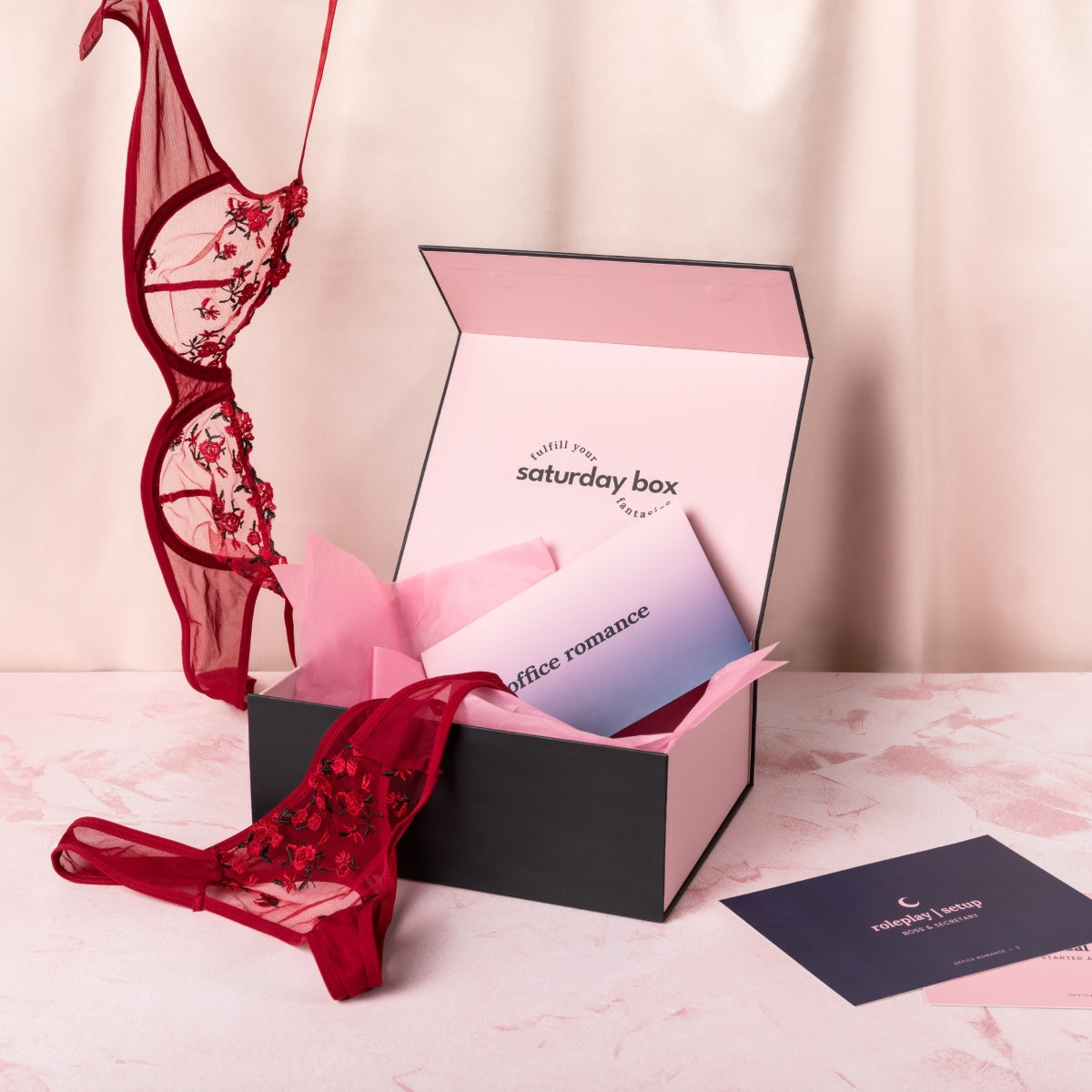 Opened Office Romance Classic Box with pink tissue paper and branded envelope inside with floral lingerie bra dangling from the top of frame and panties draped over the edge of the box. Gameplay cards are in the foreground next to the box.