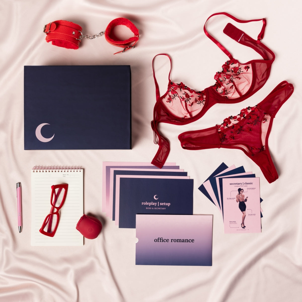 The components of the Office Romance Classic box are all laid out on a silky fabric including the magnetic box, the bright red fuzzy cuffs, the memo pad and pink pen, the bright red secretary glasses, the rose shaped suction vibrator, the red floral lingerie, and an assortment of gameplay and character cards included with the set. 