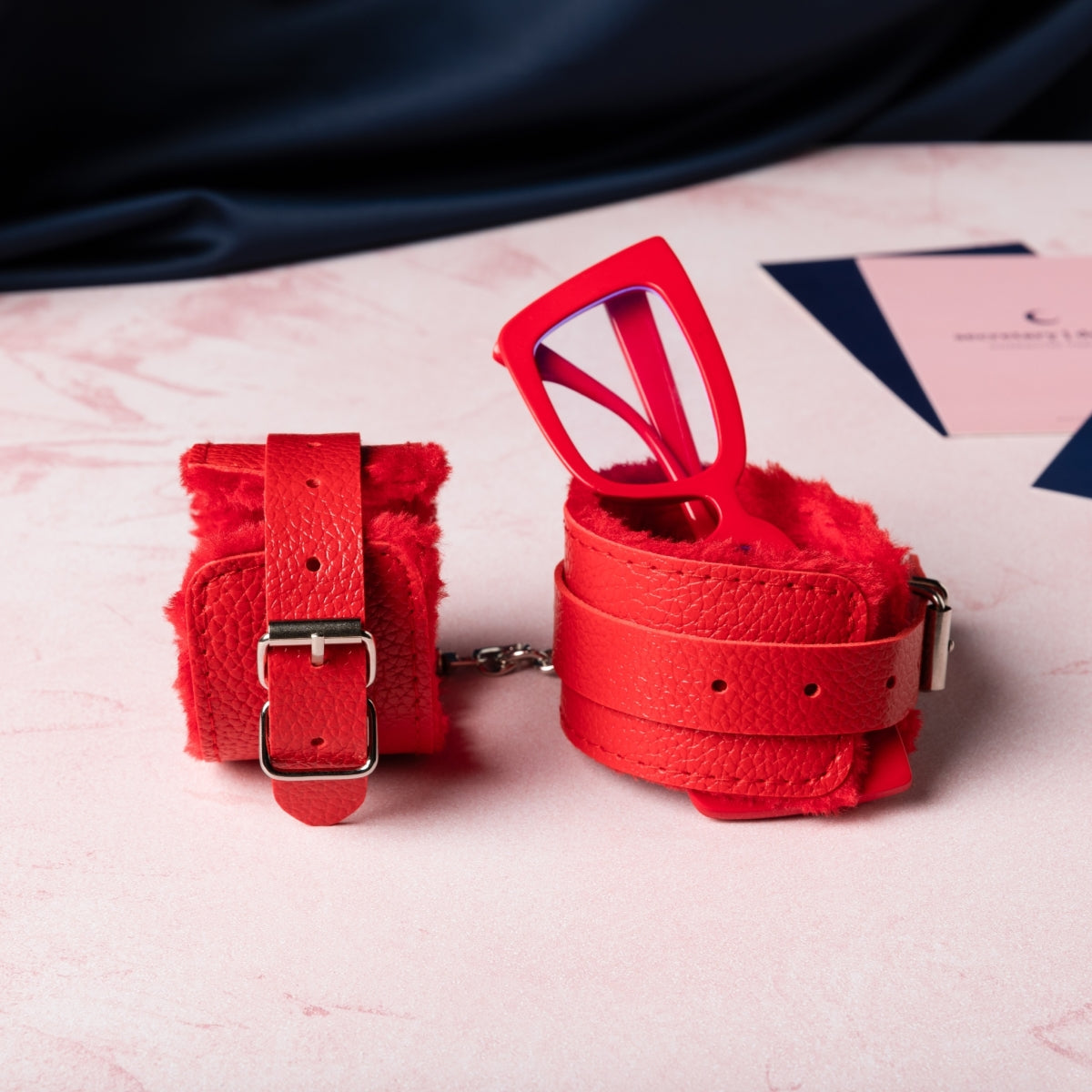 Bright red fuzzy cuffs are set out with one tipped on its side and one holding a pair of bright red glasses upright. There are Office Romance character cards in the background, out of focus