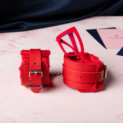Bright red fuzzy cuffs are set out with one tipped on its side and one holding a pair of bright red glasses upright. 