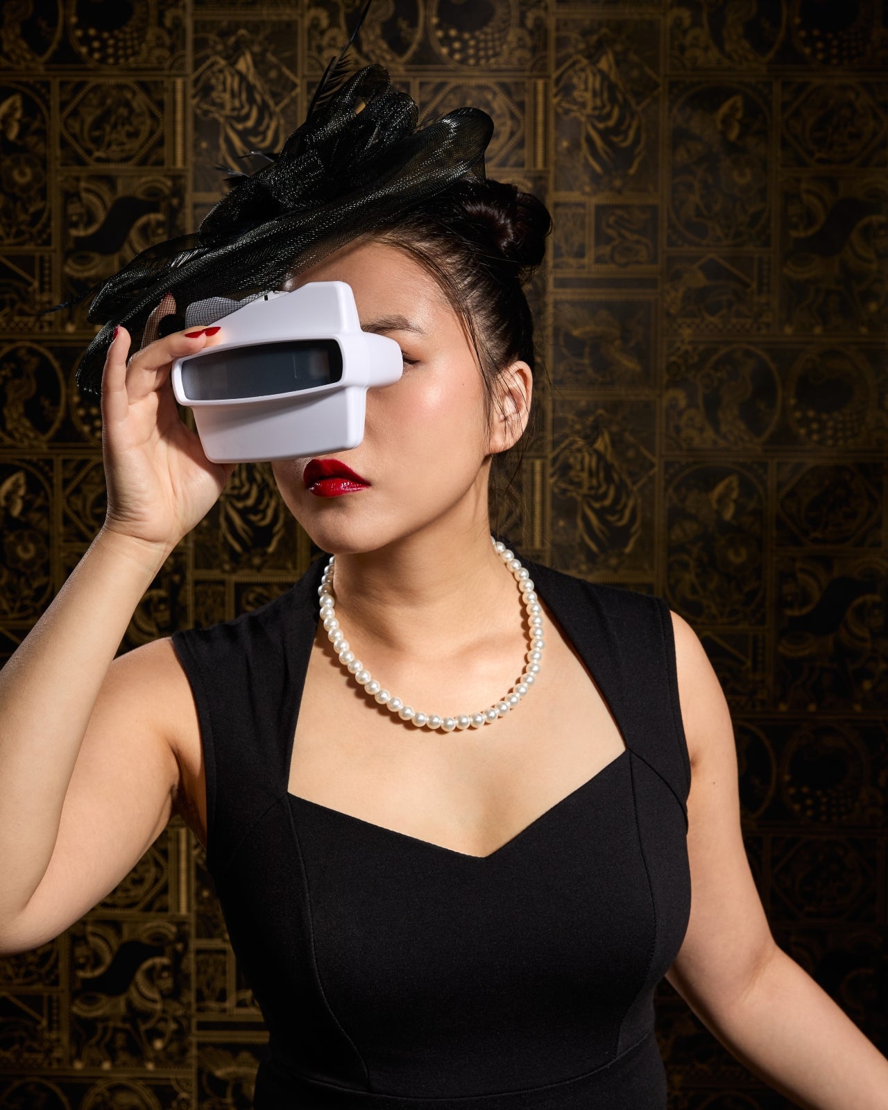 A woman weaing a fascinator and a pearl necklace looking through a white viewfinder 