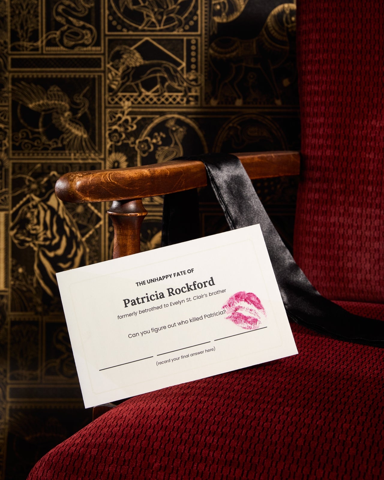 A beige envelope marked with a lipstick imprint propped up on a red upholstered chair with a strip of black fabric on the arm of the chair