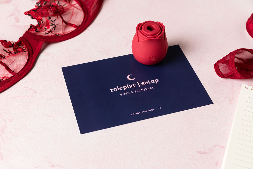 Rose suction vibrator on top of a gameplay card from the Office Romance set with red floral lingerie in the background and a memo pad peeking out in the corner