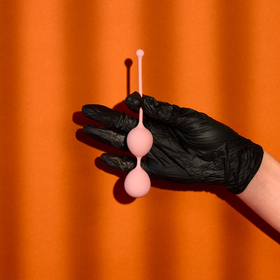 Black gloved hand holding Hard Time Ben-Wa Balls in front of orange background
